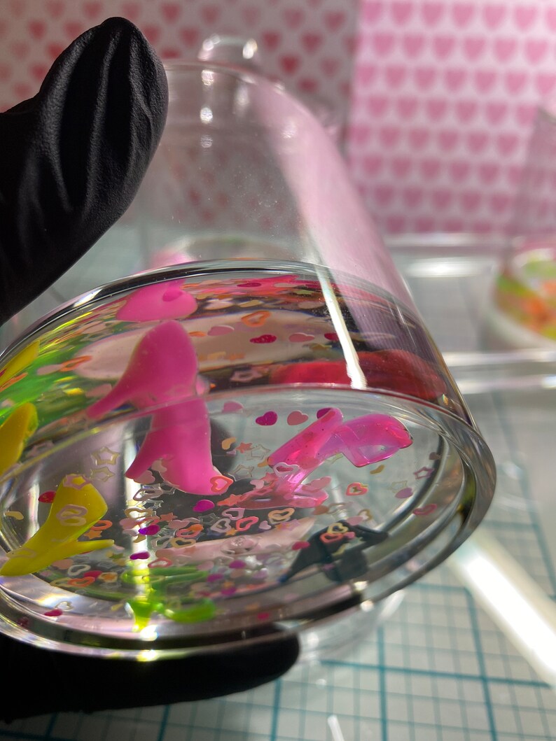 Puede incluir: Un tarro de vidrio transparente lleno de agua y purpurina de colores. La purpurina incluye estrellas y corazones rosas, amarillos, verdes y blancos. Tambi&eacute;n hay dos figuritas rosas dentro del tarro.