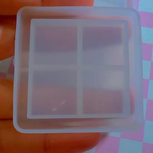 May include: A square, translucent silicone mold featuring a raised outer border and a central divider that creates four equal, smaller square compartments. The mold has a smooth, flexible texture and a clear, frosted appearance throughout its structure.