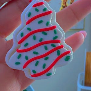 May include: A white Christmas tree-shaped ornament with red and green accents. The ornament has a red beaded chain for hanging. The tree design features red stripes and green sprinkles, resembling a decorated cookie.