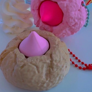 Puede incluir: Primer plano de tres objetos texturizados en tonos rosa y beige. Dos son rosas, uno con una forma central rosa y el otro con un interior rosa. El tercero es beige con un centro rosa. Un dije dice "LOVE BUG".