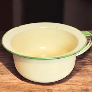 May include: A vintage, off-white enamelware bowl with a green rim and handle. The bowl is filled with a pale yellow liquid, and sits on a wooden surface. The bowl has a classic, slightly worn appearance.