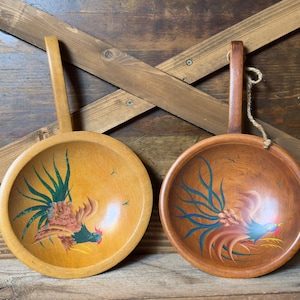 May include: Two round wooden bowls with long handles, each featuring a hand-painted rooster design. The left bowl is a lighter wood tone with a green and brown rooster, while the right bowl is a darker wood tone with a blue and brown rooster.