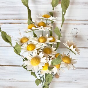 May include: A bouquet of felt daisies with white petals and yellow centers. The daisies are arranged in a vase with green stems and leaves.