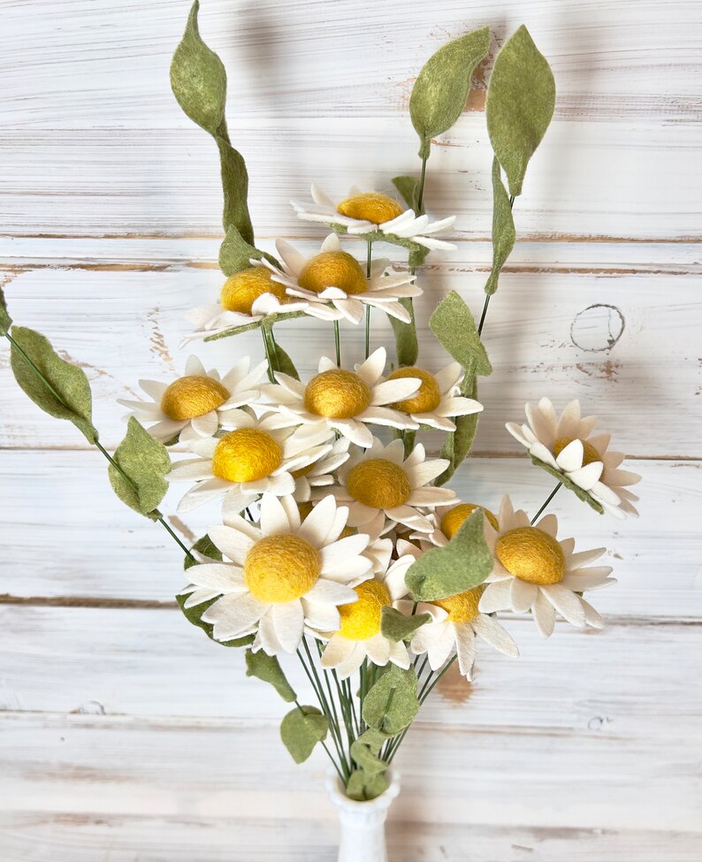 May include: A bouquet of felt daisies with white petals and yellow centers. The daisies are arranged in a vase with green stems and leaves.