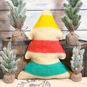 Puede incluir: Un coj&iacute;n con forma de &aacute;rbol que presenta rayas horizontales en amarillo, crema, rojo, crema y verde azulado. El coj&iacute;n tiene una textura suave similar al fieltro y est&aacute; colocado en el centro, rodeado por cuatro peque&ntilde;os pinos artificiales.