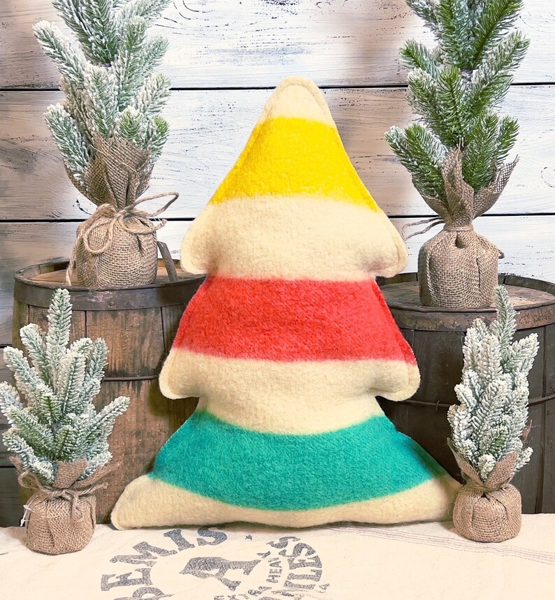 Puede incluir: Un coj&iacute;n con forma de &aacute;rbol que presenta rayas horizontales en amarillo, crema, rojo, crema y verde azulado. El coj&iacute;n tiene una textura suave similar al fieltro y est&aacute; colocado en el centro, rodeado por cuatro peque&ntilde;os pinos artificiales.