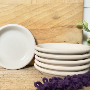 May include: A stack of six small, off-white ceramic dishes with a raised rim. One dish is separate, next to the stack. The dishes are on a wooden surface, with a wooden cutting board in the background. A sprig of greenery and purple decoration are also present.