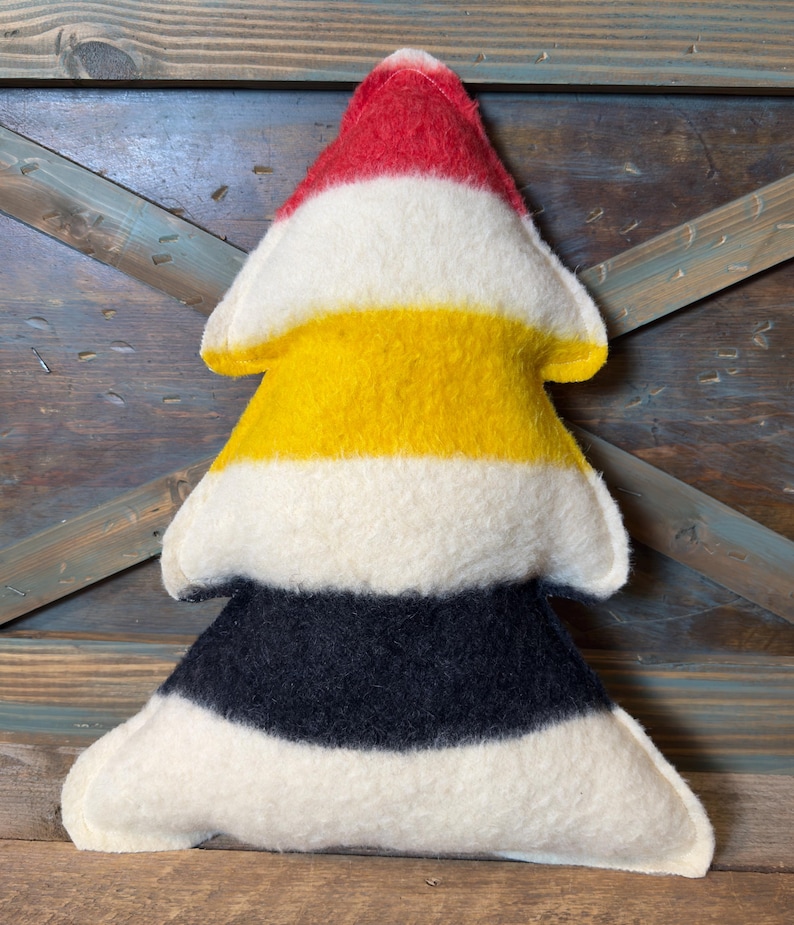 Puede incluir: Un coj&iacute;n suave con forma de &aacute;rbol que presenta franjas horizontales de fieltro en colores rojo, blanco, amarillo, blanco, negro y blanco. La decoraci&oacute;n rellena tiene una punta superior y una base ancha de tres niveles con costuras.