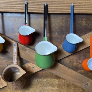 May include: A collection of six enameled metal pouring pots with long handles, featuring a variety of solid colors including yellow, red, green, blue, and orange, plus one brown patterned pot, all hanging from a wooden structure.
