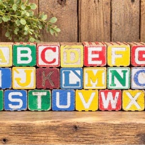 May include: A set of twenty-six wooden alphabet blocks arranged in three rows. Each cube features a painted letter in a contrasting color, including red, blue, green, and yellow. The blocks show signs of wear with visible paint chipping.