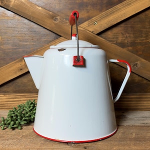 May include: White enamelware coffee pot with red trim and handle accents. The vintage kettle features a lid and a spout. The handle is red and black. The pot is set against a wooden background.