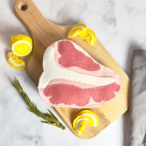 May include: A felt toy steak on a wooden cutting board with lemon slices and a sprig of rosemary.
