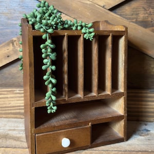 May include: A brown wooden desk organizer featuring five vertical slots for documents, a horizontal shelf, and a single drawer with a small white knob. A trailing green artificial succulent plant hangs over the top edge of the organizer.