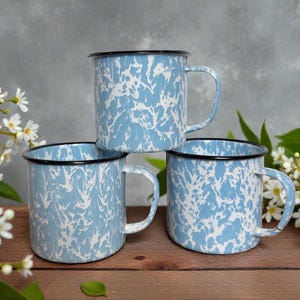 May include: Three blue and white enamel mugs with black rims and handles are stacked on a wooden surface. The mugs have a speckled pattern and are surrounded by white flowers and green leaves. The mugs are a classic camping style.