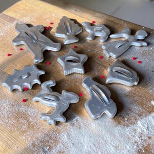 May include: A collection of nine silver-colored metal cookie cutters in various holiday shapes, including a star, a bell, a tree, an angel, and a gingerbread person, arranged on a wooden surface with scattered red sprinkles and white flour.