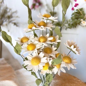 May include: A bouquet of felt daisies with yellow centers and white petals. The daisies are arranged in a vase with green stems and leaves.