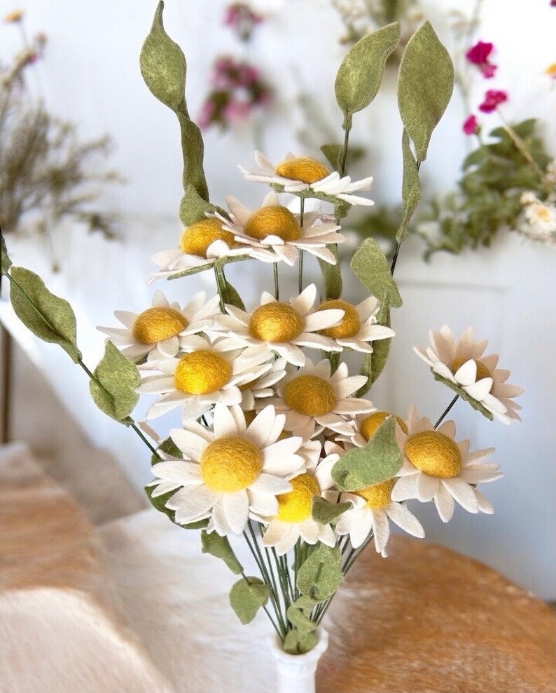 May include: A bouquet of felt daisies with yellow centers and white petals. The daisies are arranged in a vase with green stems and leaves.