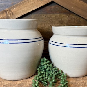 Puede incluir: Dos vasijas de cerámica de color blanco roto con una boca ancha y un cuerpo redondeado. Cada recipiente presenta dos finas rayas horizontales de color azul marino que rodean la parte superior del exterior. Las vasijas tienen un acabado liso.