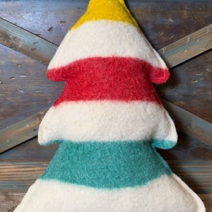 Puede incluir: Un coj&iacute;n suave con forma de &aacute;rbol que presenta rayas horizontales en amarillo, blanco, rojo, blanco, verde azulado y blanco. La tela tiene una textura afieltrada y difusa, con costuras visibles a lo largo de los bordes.