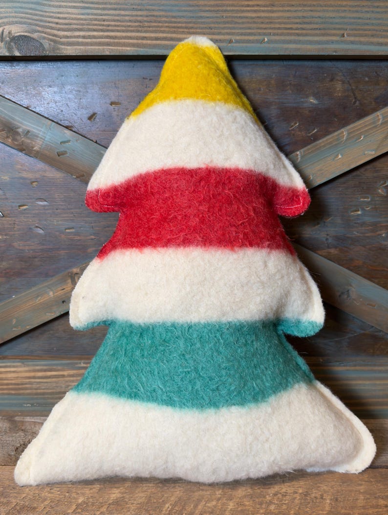 Puede incluir: Un coj&iacute;n suave con forma de &aacute;rbol que presenta rayas horizontales en amarillo, blanco, rojo, blanco, verde azulado y blanco. La tela tiene una textura afieltrada y difusa, con costuras visibles a lo largo de los bordes.
