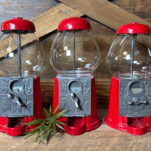 May include: Three red gumball machines with clear glass globes and silver-toned metal coin mechanisms featuring embossed leaf patterns. Each machine has a round red lid, a central metal rod, and a small dispensing tray at the base.