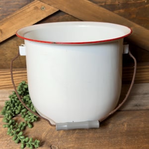 May include: White enamel bucket with a red rim and a brown handle. The bucket has a gray handle grip and is sitting on a wooden surface. A green plant is in front of the bucket.