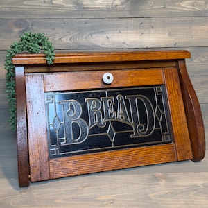 May include: A wooden bread box with a dark brown finish, featuring a front-hinged door with a central white ceramic knob. The door contains a black glass panel with the word BREAD etched in a decorative font and geometric leaded lines.
