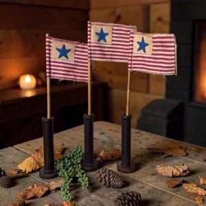 May include: Three decorative flags with red and white stripes and a blue star on a tan background. The flags are mounted on wooden dowels and set in black holders. The flags are arranged on a rustic wooden table with leaves and pinecones.