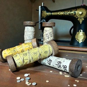 May include: A collection of wooden spools wrapped with vintage measuring tapes, in yellow and white, with black numbering. The spools are arranged on a wooden surface, with a vintage sewing machine in the background.