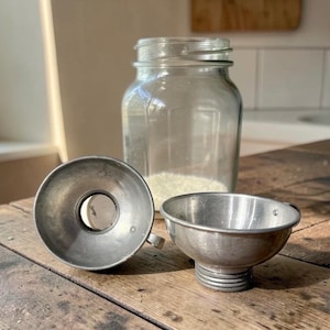 May include: A vintage kitchen scene featuring a clear glass jar, two metal funnels, and a wooden surface. The funnels are silver-toned, one with a handle and the other with a ribbed base. The jar is partially filled with a white substance.