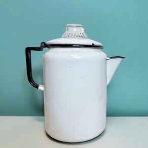 May include: White enamel coffee pot with a black handle and rim. The pot has a clear glass knob on the lid and a spout for pouring. The background is a solid teal color.