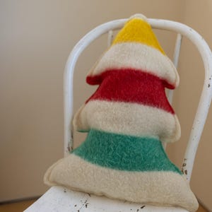 Puede incluir: Un coj&iacute;n de fieltro con forma de &aacute;rbol que presenta rayas horizontales en amarillo, rojo, crema y verde. El objeto textil suave y texturizado tiene una parte superior puntiaguda y una base ancha, con costuras visibles en los bordes.