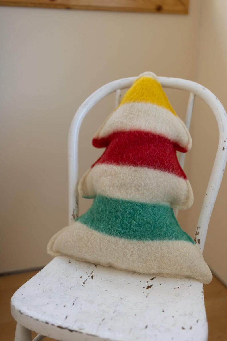 Puede incluir: Un coj&iacute;n de fieltro con forma de &aacute;rbol que presenta rayas horizontales en amarillo, rojo, crema y verde. El objeto textil suave y texturizado tiene una parte superior puntiaguda y una base ancha, con costuras visibles en los bordes.