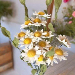May include: A bouquet of felt daisies with white petals and yellow centers. The stems are green and the flowers are arranged in a vase.