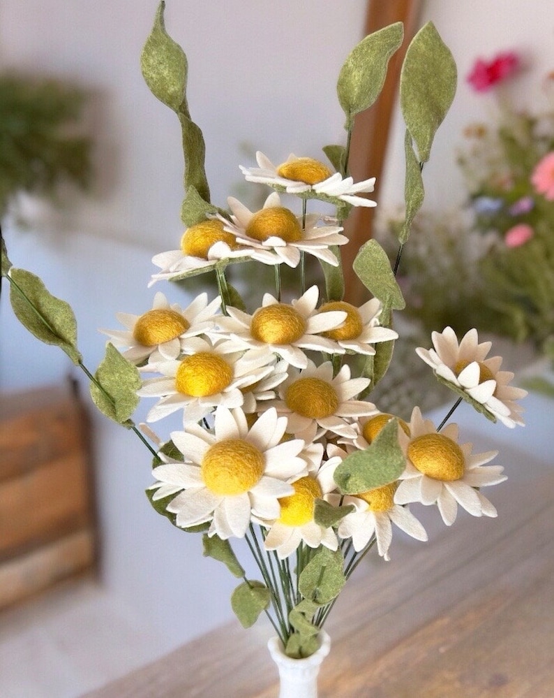 May include: A bouquet of felt daisies with white petals and yellow centers. The stems are green and the flowers are arranged in a vase.
