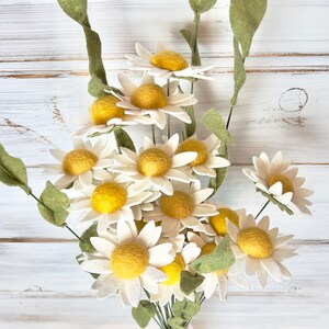 May include: A bouquet of felt daisies in a white vase. The daisies have white petals and yellow centers. The stems are green.