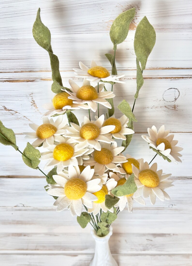 May include: A bouquet of felt daisies in a white vase. The daisies have white petals and yellow centers. The stems are green.