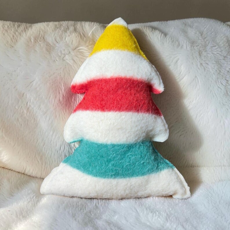 Puede incluir: Un coj&iacute;n de peluche con forma de &aacute;rbol que presenta rayas horizontales en amarillo, blanco, rojo, blanco, verde azulado y blanco. La tela suave y difusa crea una apariencia texturizada en toda la superficie del objeto.