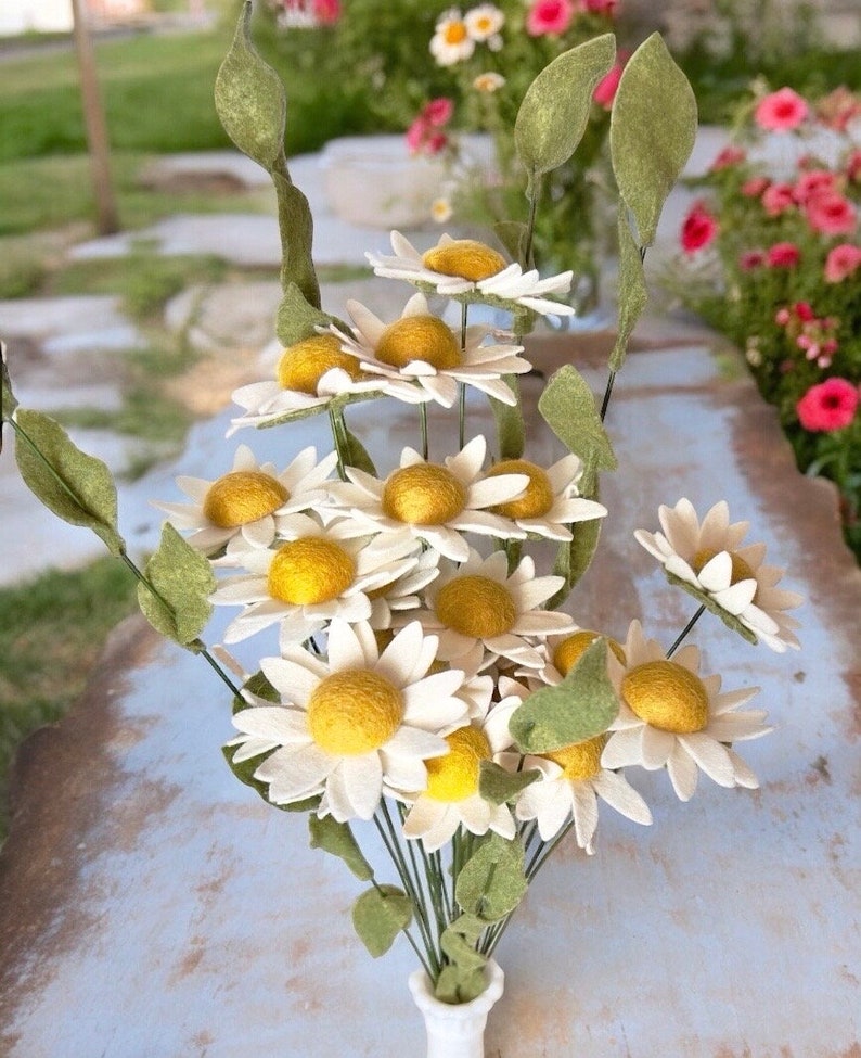 May include: A bouquet of felt daisies in shades of white and yellow with green stems. The daisies are arranged in a vase.