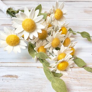 May include: A bouquet of felt daisies with white petals and yellow centers. The daisies are arranged in a bunch with green felt leaves.