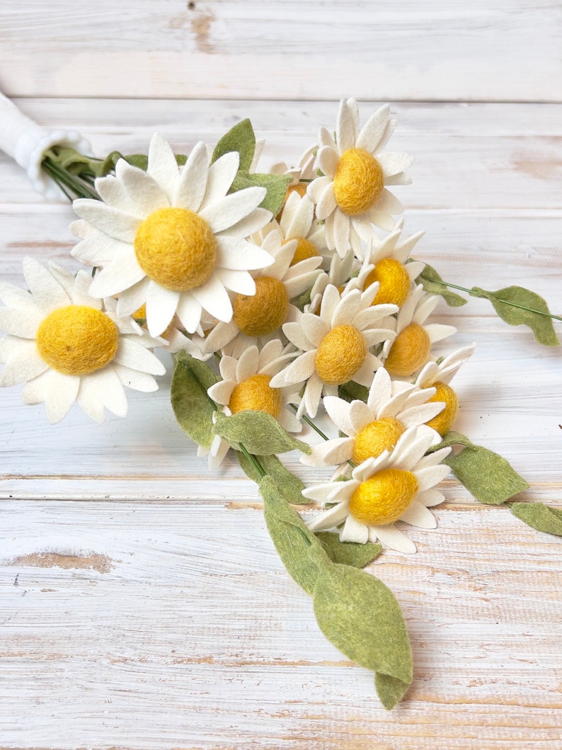 May include: A bouquet of felt daisies with white petals and yellow centers. The daisies are arranged in a bunch with green felt leaves.