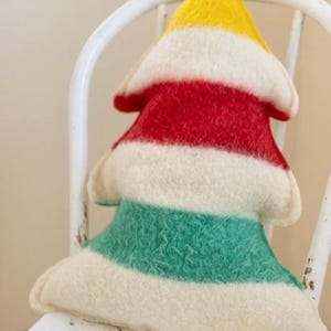 Puede incluir: Un coj&iacute;n con forma de &aacute;rbol hecho de lana fieltrada. El coj&iacute;n presenta rayas horizontales en amarillo, rojo, crema y verde. La textura de la lana es visible en toda la superficie del coj&iacute;n, que tiene un aspecto suave, redondeado y acolchado.