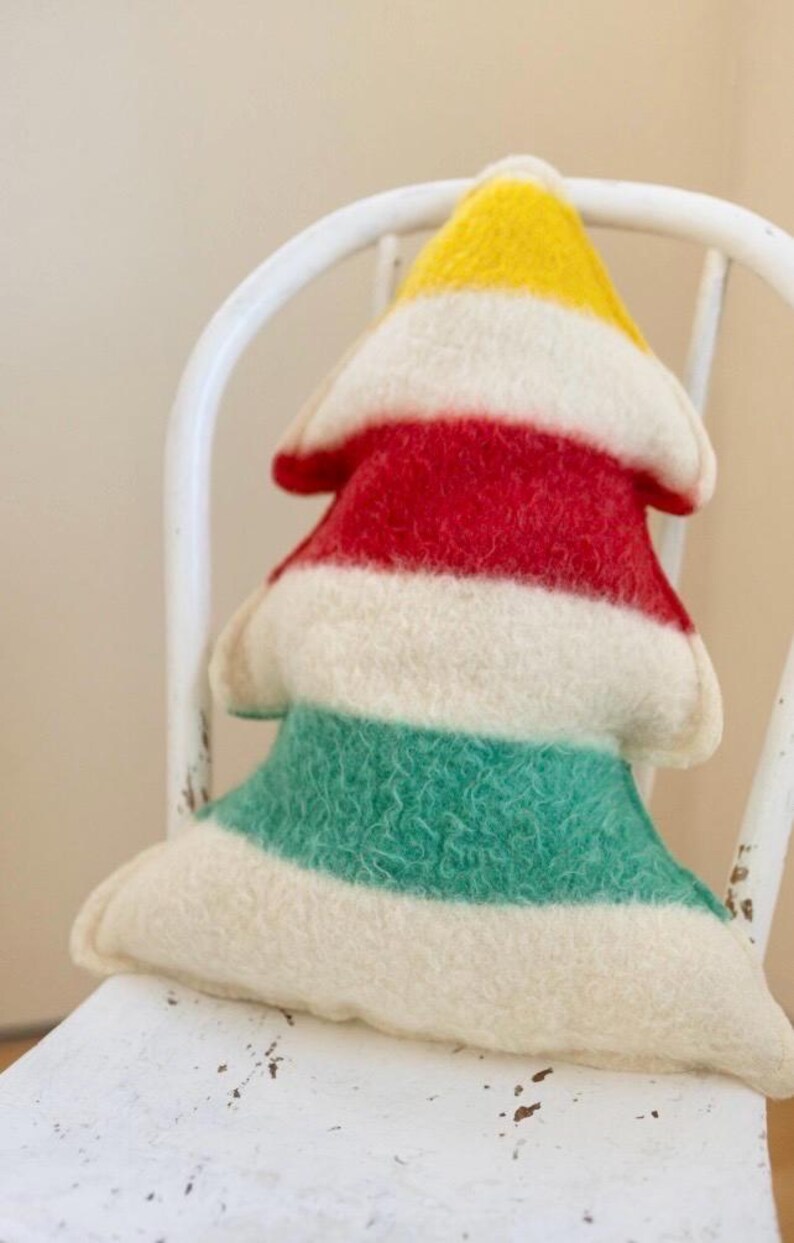 Puede incluir: Un coj&iacute;n con forma de &aacute;rbol hecho de lana fieltrada. El coj&iacute;n presenta rayas horizontales en amarillo, rojo, crema y verde. La textura de la lana es visible en toda la superficie del coj&iacute;n, que tiene un aspecto suave, redondeado y acolchado.
