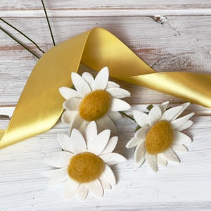 May include: Three white felt daisies with yellow centers are arranged on a white wooden surface with a yellow satin ribbon.