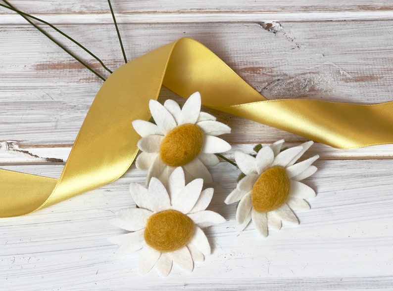May include: Three white felt daisies with yellow centers are arranged on a white wooden surface with a yellow satin ribbon.