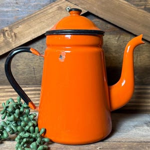 May include: An orange enamelware coffee pot with a black handle and rim. The vintage pot has a lid and a spout. A sprig of green faux succulents is in the foreground. The pot is on a wooden surface.