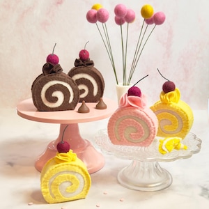 May include: Three felt cake rolls on cake stands. The cake rolls are decorated with cherries and frosting. The cake rolls are yellow, pink, and brown. The cake stands are pink and clear.