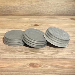 May include: Three gray metal jar lids are arranged on a wooden surface. The lids are round with a ribbed design around the edges. The background is a wooden surface with a natural wood grain.