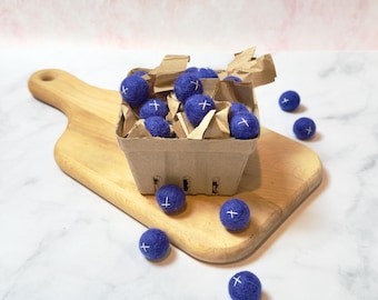 Pode incluir: Um recipiente de cartão cheio de bolas de feltro azuis com sinais de mais brancos nelas. O recipiente está sentado em uma tábua de cortar de madeira. Há várias bolas de feltro azuis espalhadas ao redor do recipiente.