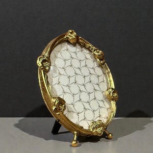 May include: An oval, gold-colored picture frame with a decorative floral design. The frame has a stand and holds a patterned, off-white insert. The frame is approximately 5 inches tall.